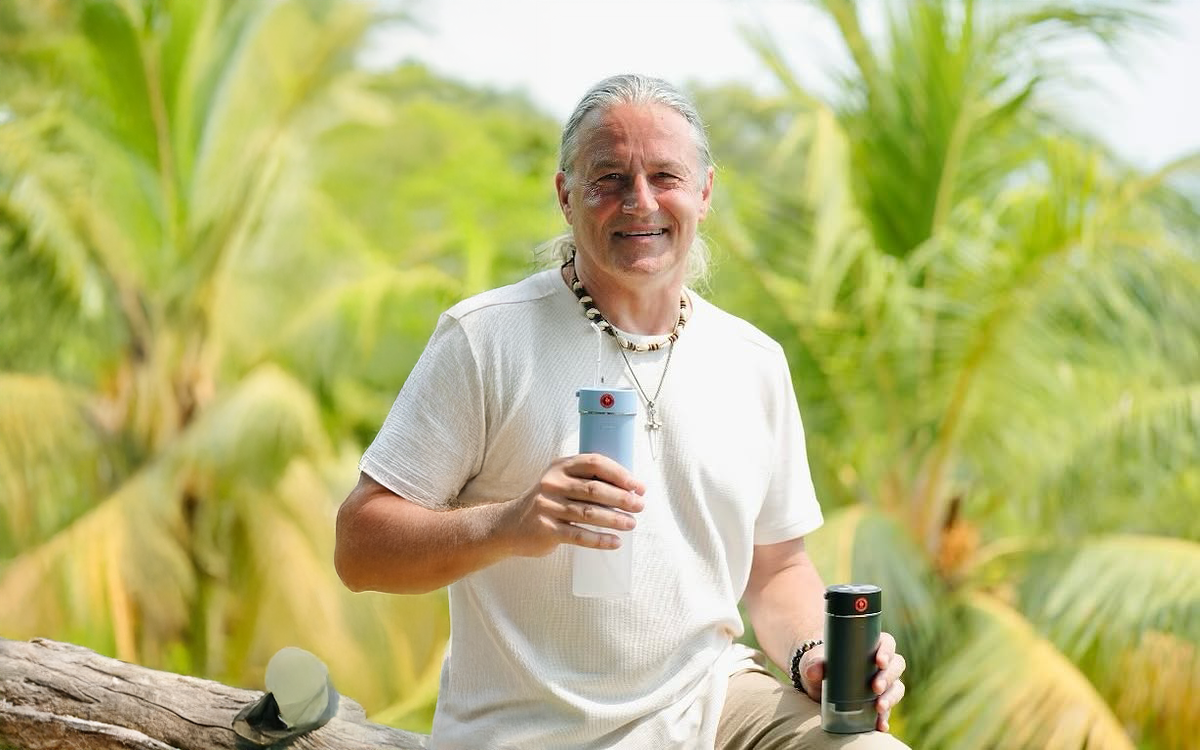 Marc the Dental Shaman smiling outdoors in a tropical setting, holding two ozone water flossers, embodying a holistic approach to oral health and wellness.