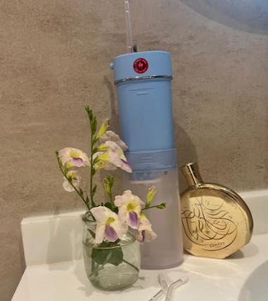 Blue ozone water flosser displayed on a bathroom counter beside a small glass vase with flowers and a round gold perfume bottle, creating a calm and elegant wellness scene.