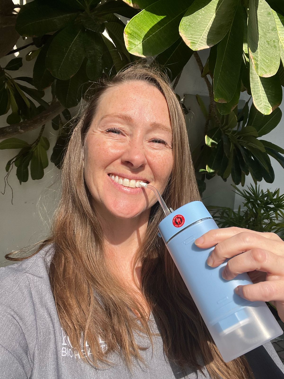 Smiling woman holding a blue ozone water flosser outdoors, standing in front of lush green foliage, promoting natural oral care and wellness.