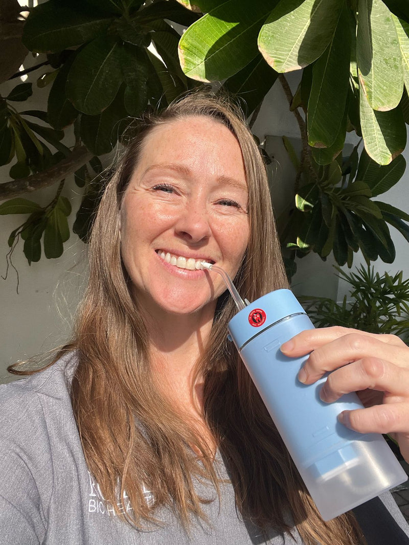 Smiling woman holding a blue ozone water flosser outdoors, standing in front of lush green foliage, promoting natural oral care and wellness.