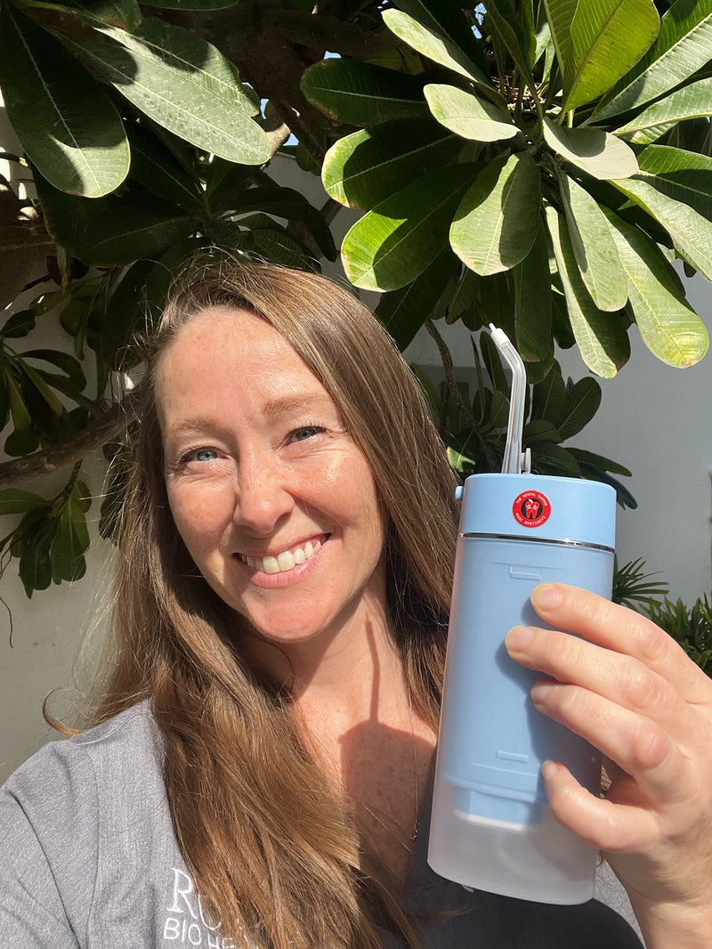 Smiling woman holding a blue ozone water flosser outdoors, standing in front of lush green foliage, promoting natural oral care and wellness.