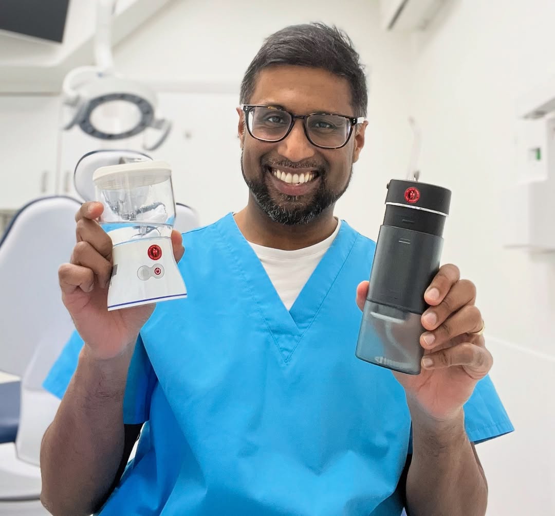Smiling dentist in blue scrubs holding two ozone water flossers inside a modern dental clinic, demonstrating advanced tools for holistic oral care.